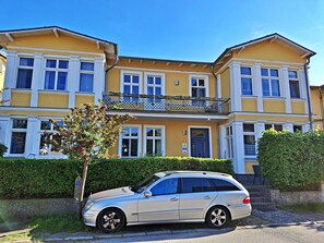 Un immeuble de deux étages avec un balcon, une voiture garée et une haie devant.