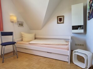 A small, single-bed room with a chair, a bedside table, a wall-mounted shelf, and a picture frame.