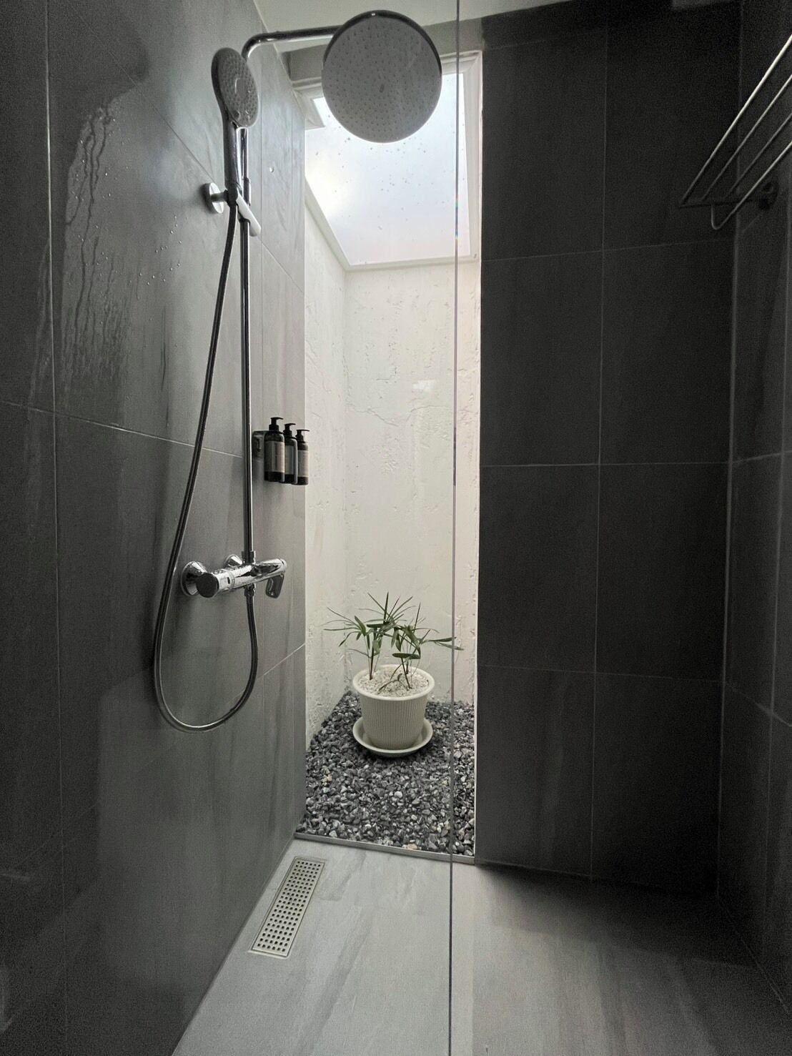 Een moderne douche met een handdouche en een muurbeugel. In de hoek staat een kleine potplant op een grindbed.