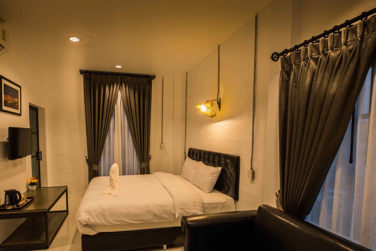 A hotel room with a bed, a bedside table with a kettle, a chair, a TV, and a window with curtains.