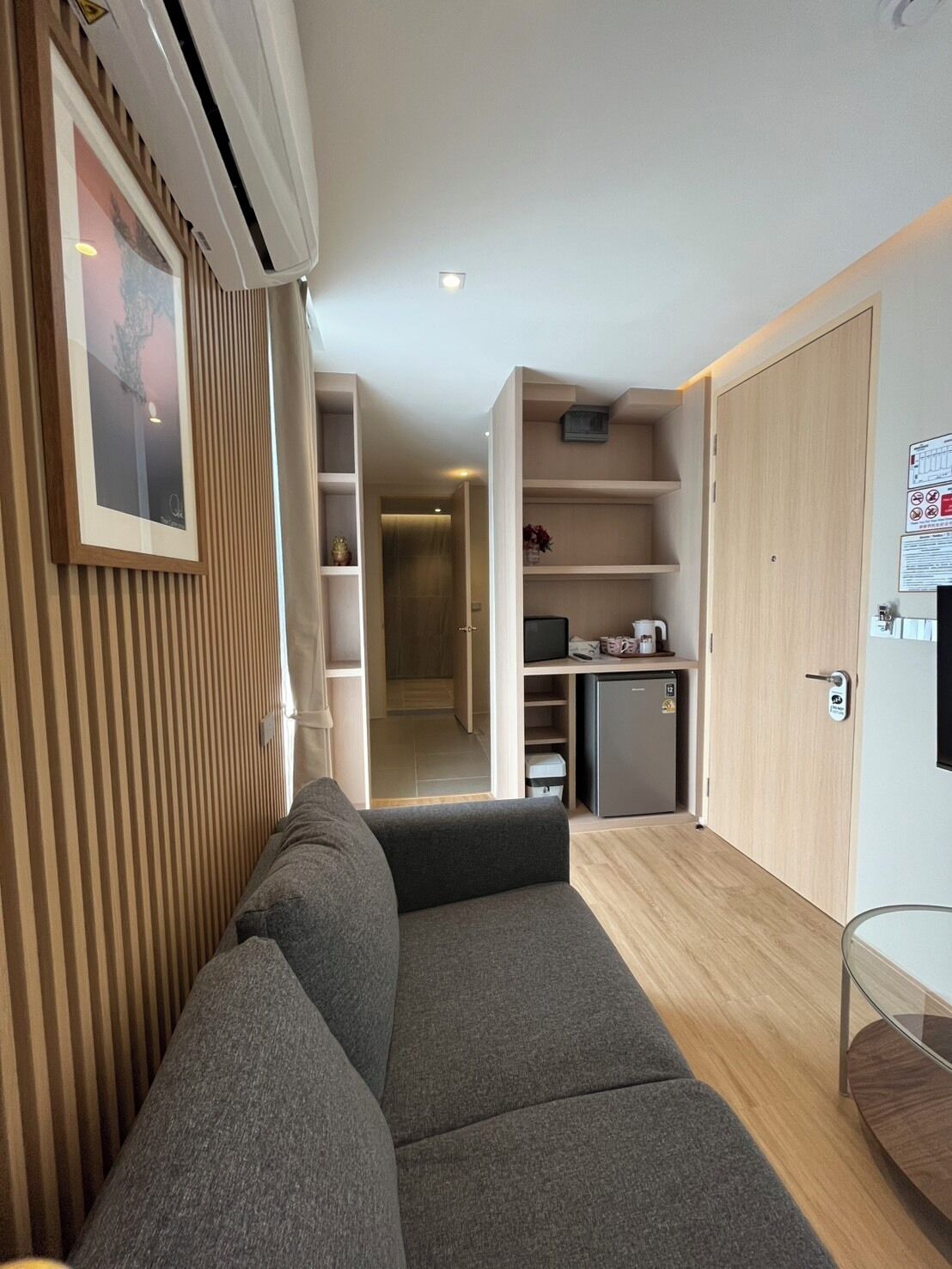 A modern living room with a grey sofa, a small kitchen area with a refrigerator and a coffee maker, and a wooden door with a lock.
