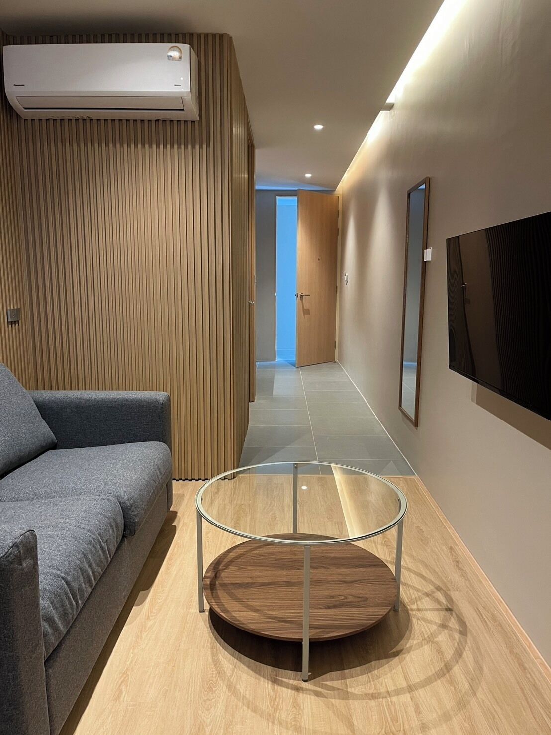 Un pasillo moderno con un panel de pared de madera, un sofá gris, una mesa redonda con superficie de vidrio y un televisor montado.