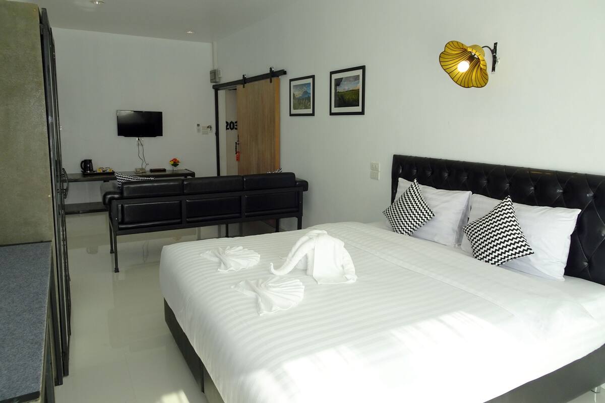 A hotel room with a large bed, a desk, a chair, a TV, and a wall-mounted light fixture.