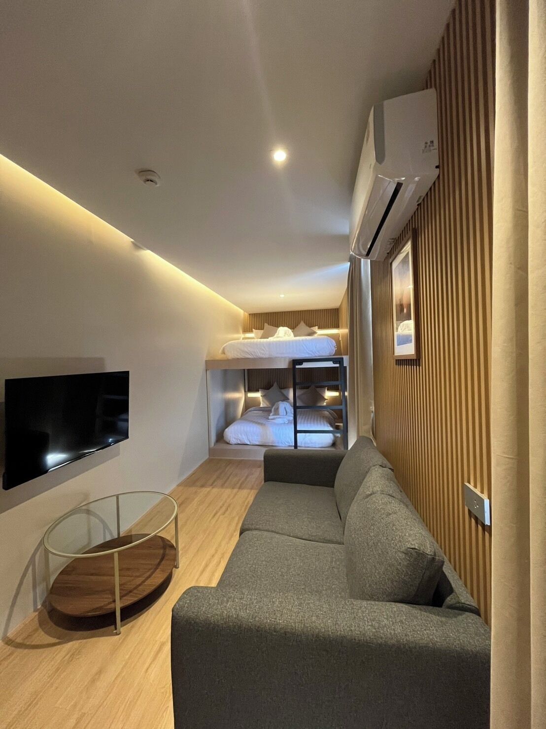 A modern hotel room with a sofa, a TV, a bunk bed, and a wooden panel wall.