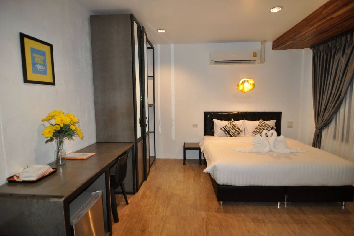 A hotel room with a bed, a desk, a chair, a vase with flowers, an air conditioning unit, and a wall-mounted light fixture.
