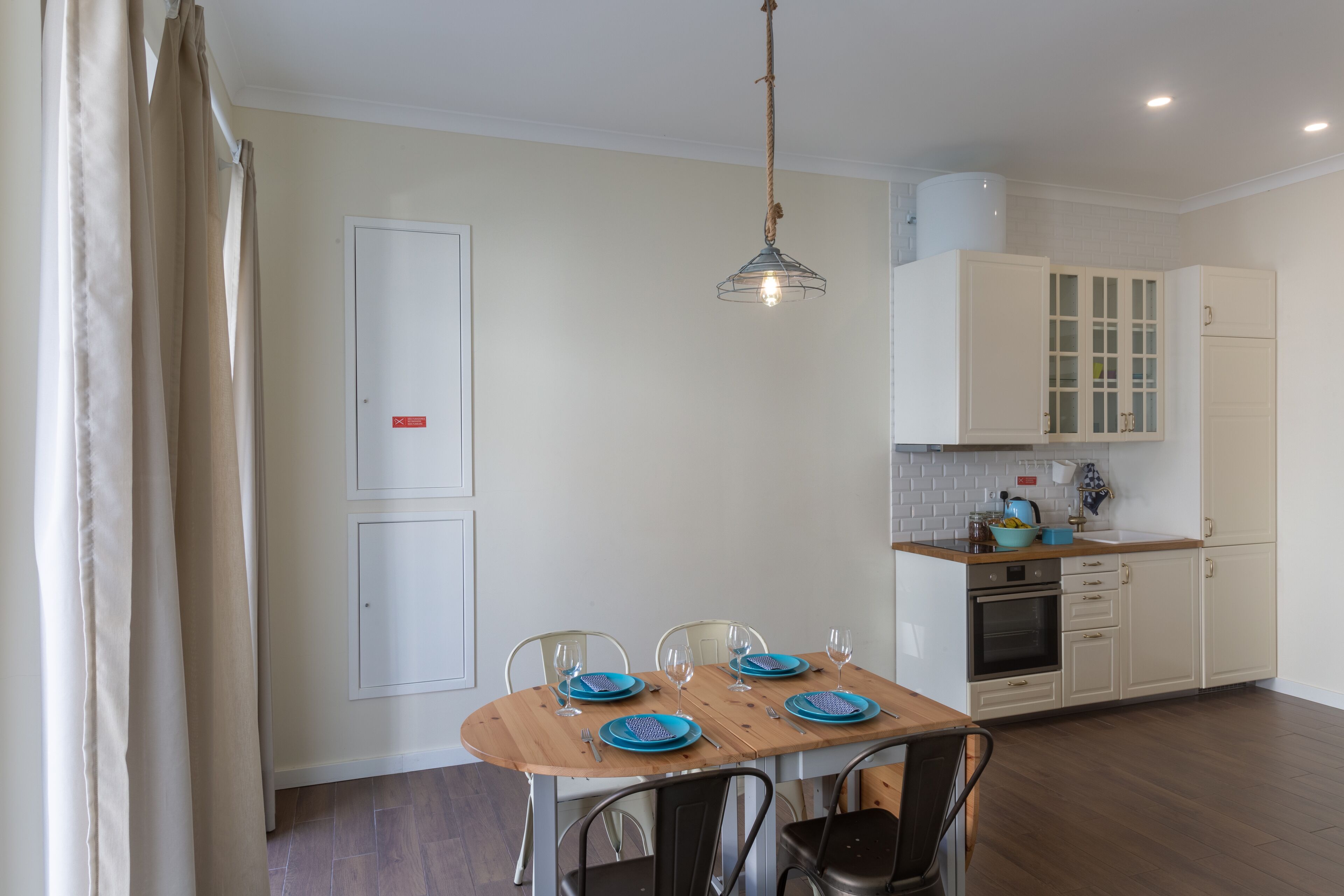 Uma sala de jantar com mesa de madeira posta para quatro pessoas, cozinha com armários brancos e luminária pendente.