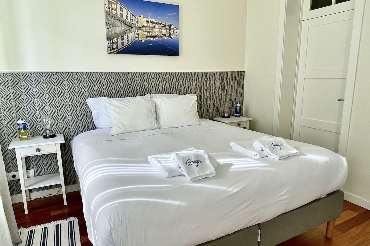 A neatly made bed with white linens and towels, a bedside table with a bottle and a glass, and a patterned wall.