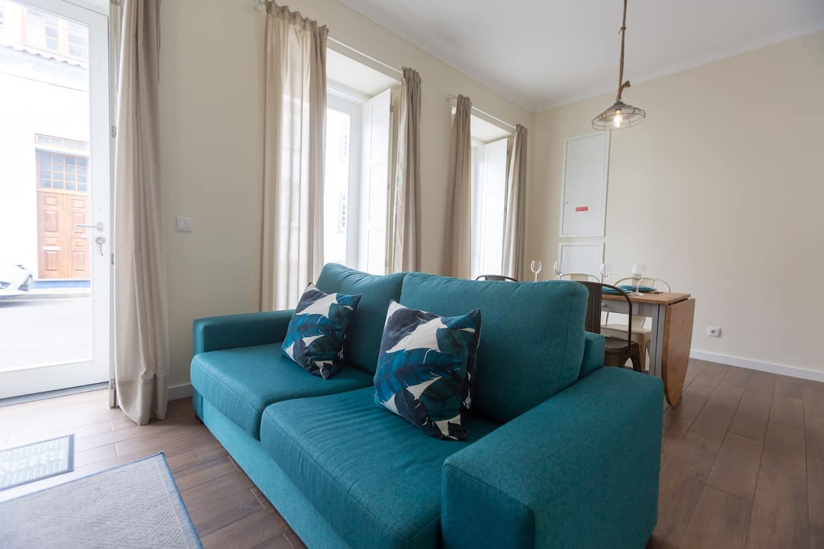 A teal sofa with patterned cushions, a wooden dining table, and a hanging light fixture in a room with large windows and a view of the street.