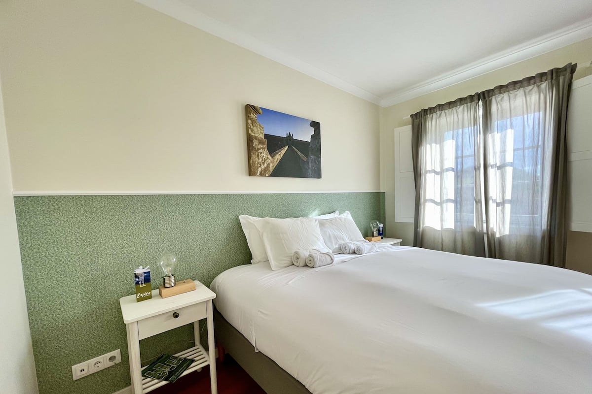 A hotel room with a large bed, a nightstand with a lamp, a painting on the wall, and a window with curtains.