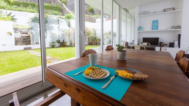 A modern dining area with a wooden table, a blue placemat, and breakfast items like waffles, toast, and fruit. - My Angra Charming House (Angra do Heroismo)