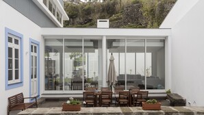A modern outdoor seating area with a large glass wall, wooden furniture, and potted plants. - My Angra Charming House (Angra do Heroismo)