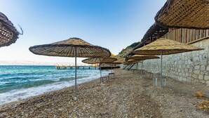 Une plage de galets bordée de parasols en chaume et d’un mur de pierres.