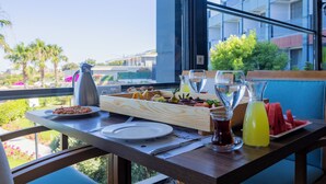 Une table à manger dressée avec divers plats, une carafe de thé et un verre d’une boisson jaune, le tout mis en valeur par la vue d’un bâtiment moderne et de palmiers.