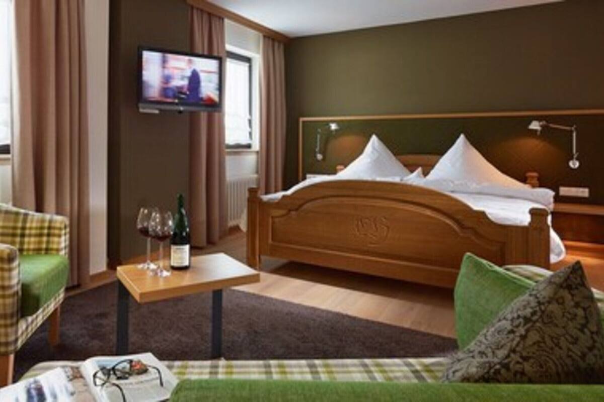 A hotel room with a bed, a television, a chair, a small table with a bottle and glasses, and a green sofa.
