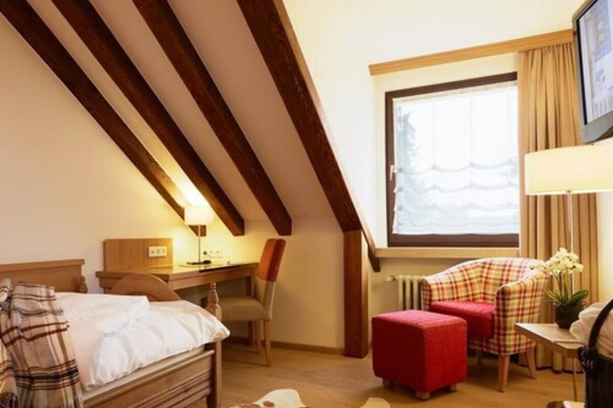 A cozy hotel room with a wooden ceiling, a bed with a plaid blanket, a desk with a lamp, a chair, a red ottoman, and a window with curtains.
