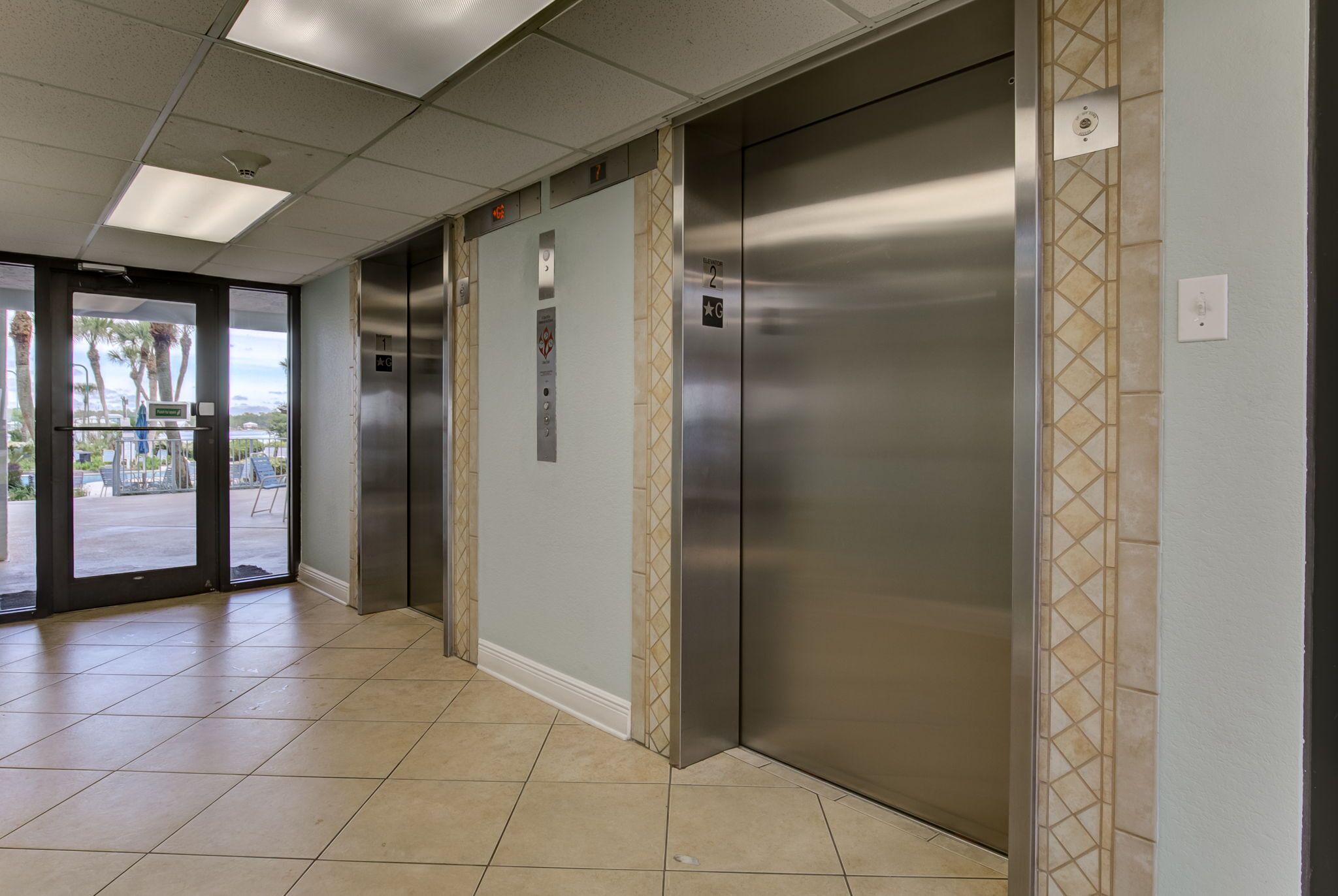 Et rent og moderne elevatormiljø med rustfrie elevatorer, flisegulv og udsigt udendørs gennem glasdøre.