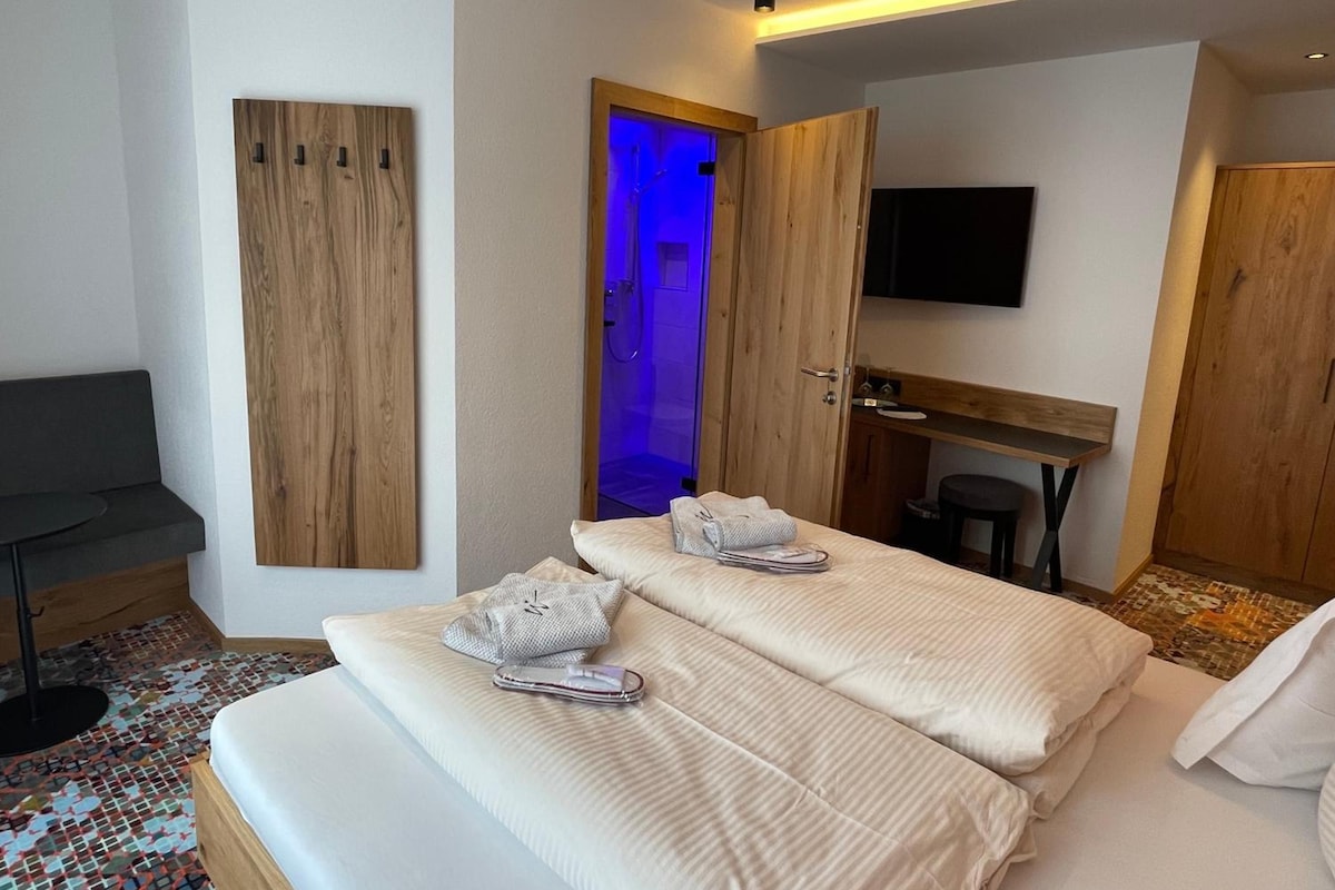 A hotel room with a bed, a desk, a chair, a TV, and a shower with blue lighting.
