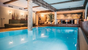 Een binnenzwembad met een helderblauw wateroppervlak, houten balken in het plafond en een zithoek met lounge stoelen en een potplante.'