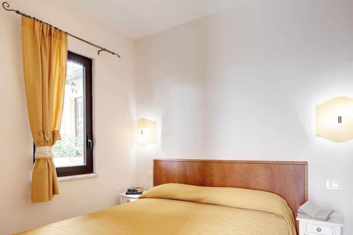A bedroom with a wooden headboard, a bed with a beige blanket, a nightstand with a lamp, and a window with curtains.