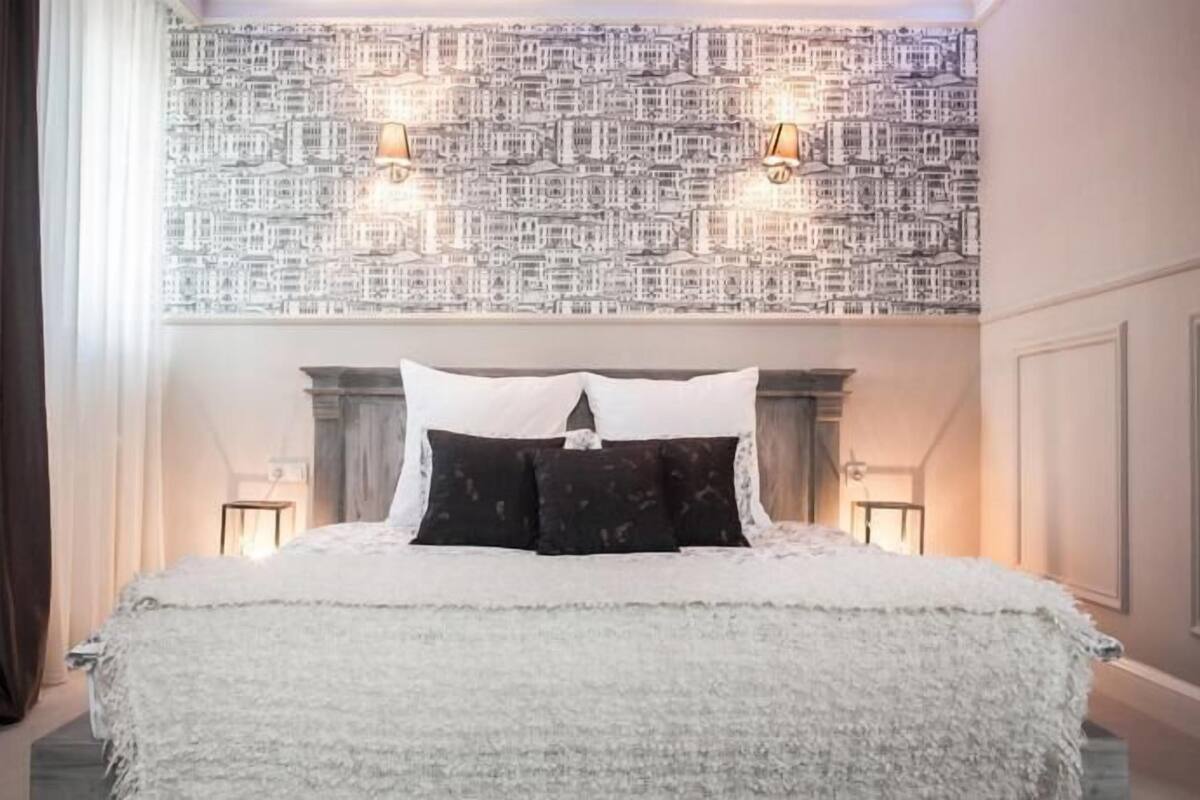A bedroom with a large bed, a patterned wallpaper, two wall-mounted lamps, and a window with curtains.