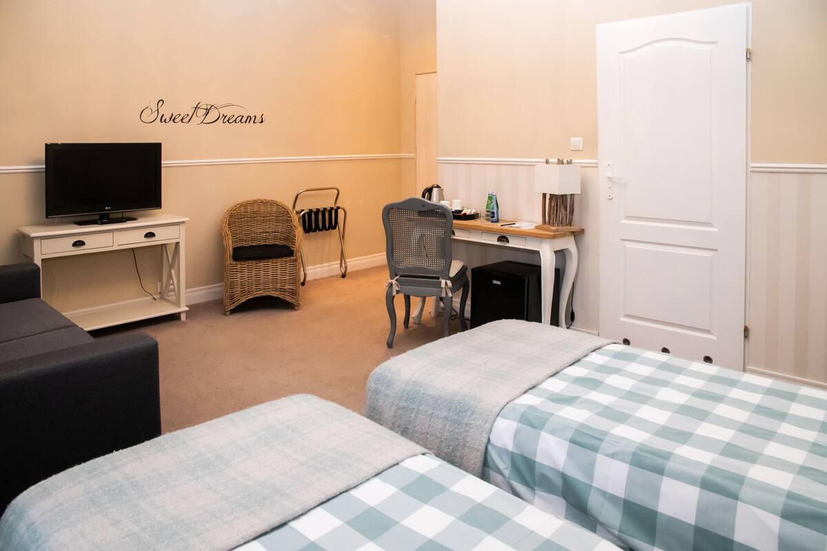 A hotel room with two beds, a television, a desk with a computer, a chair, and a wicker chair.