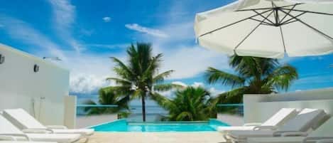 A poolside area with lounge chairs and a large umbrella, surrounded by palm trees and a clear blue sky.