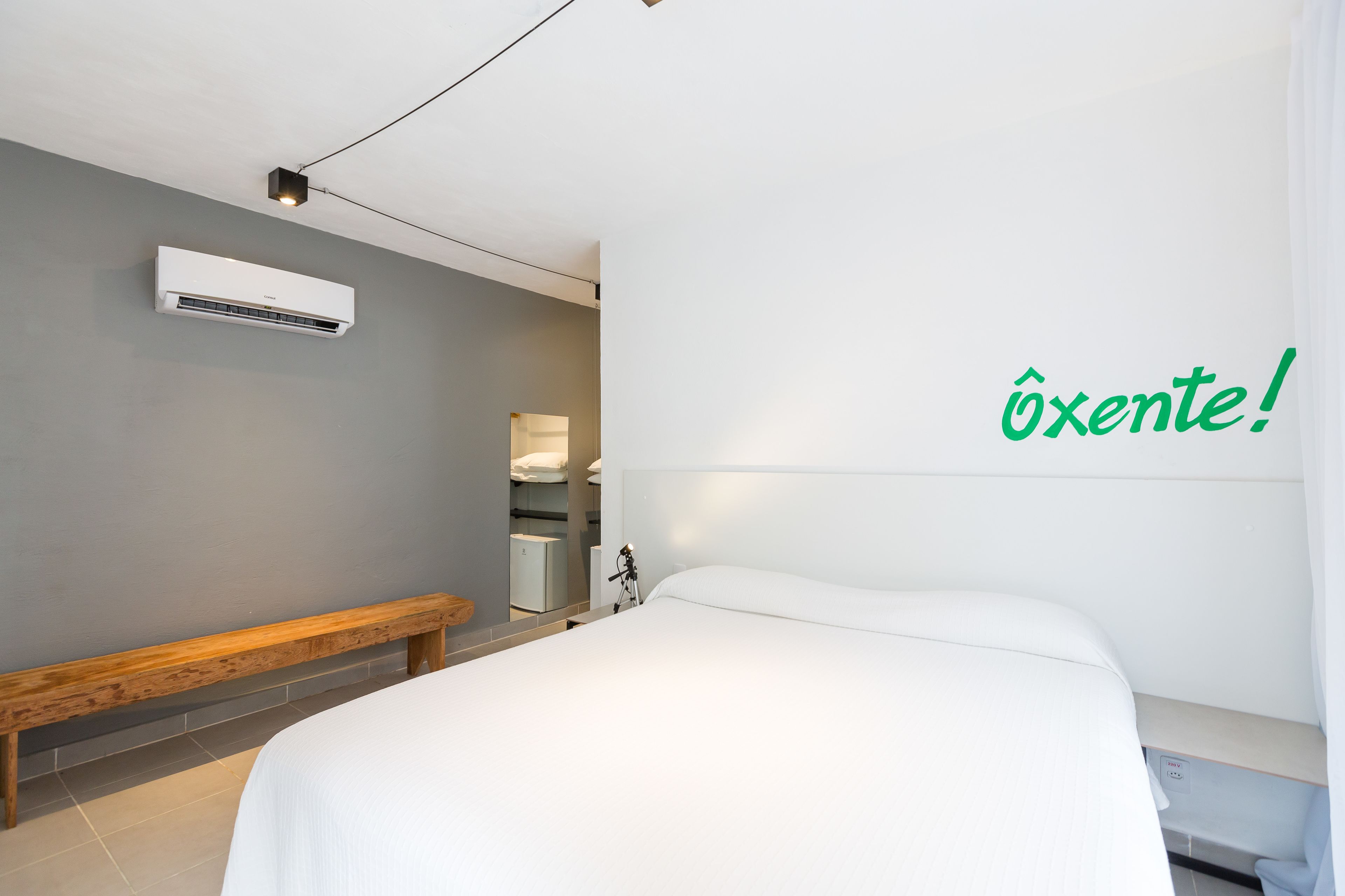 A modern hotel room with a bed, a bench, an air conditioner, and a wall-mounted light.