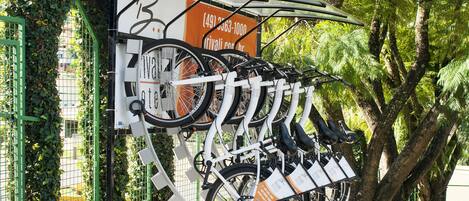 A bike rack with multiple bicycles parked, surrounded by greenery and a fence.