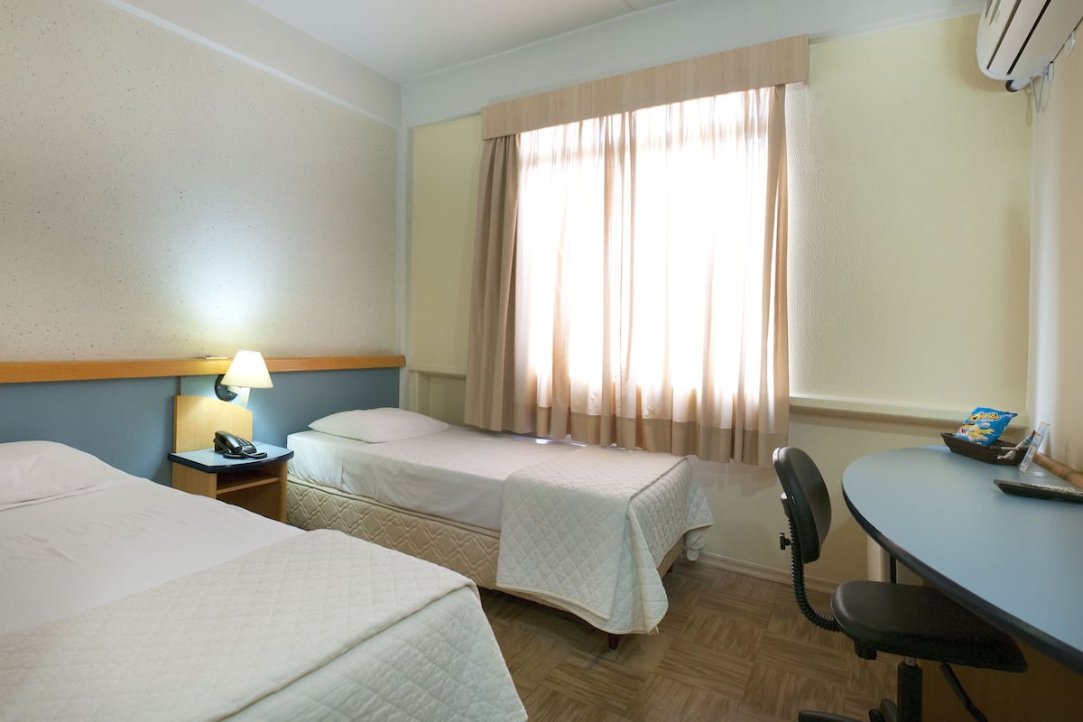 Hotellihuone, jossa on kaksi sänkyä, työpöytä, tuoli, lamppu ja ikkuna, jossa on verhot.