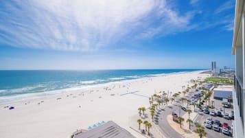 Blick auf einen Strand mit Parkplatz, Palmen und einem Gebäude im Hintergrund.