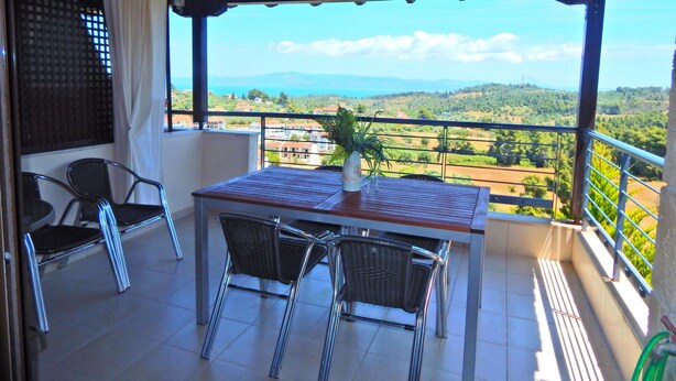Un balcón con una mesa y sillas, una planta en maceta y vistas a un paisaje con edificios y montañas.