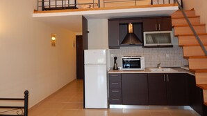 Una cocina compacta con horno empotrado, microondas y refrigerador. Presenta armarios oscuros, una escalera de madera y un pequeño comedor con una silla.