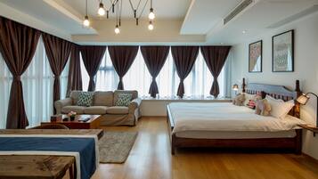 A spacious hotel room with a large bed, a sofa, a wooden table, and a desk. There are multiple windows with curtains, a ceiling light fixture, and wall art.