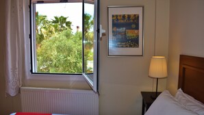 A hotel room with a bed, a window with an open view of greenery, a framed picture on the wall, and a bedside lamp.