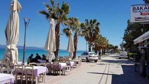 A beachfront dining area with tables and umbrellas, a parked car, and a sign for 'Bakalis Fish Restaurant' open all year.