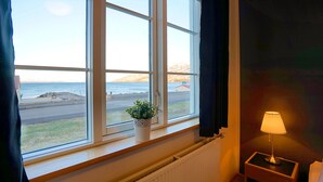 A room with a large window overlooking a beach, a small table with a lamp, and a potted plant on the windowsill. - Grenivík Guesthouse (Grenivík)