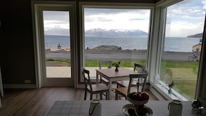 A room with a table and chairs, a view of a beach and mountains, and a door with an exit sign. - Grenivík Guesthouse (Grenivík)