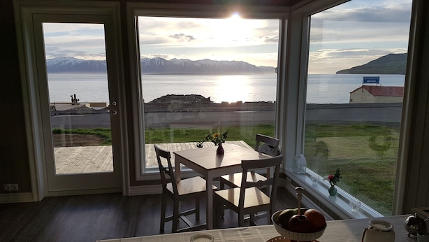 A room with a large window offering a view of a body of water and mountains. There is a table with chairs and a fruit bowl. - Grenivík Guesthouse (Grenivík)