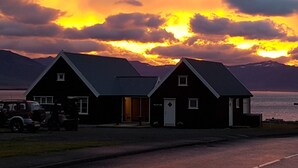 A two-story building with a dark roof and white trim, located near a body of water with mountains in the background. - Grenivík Guesthouse (Grenivík)