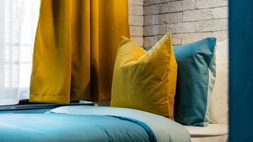 A bed with blue bedding and two pillows, one yellow and one teal, in a room with a brick wall and yellow curtains.