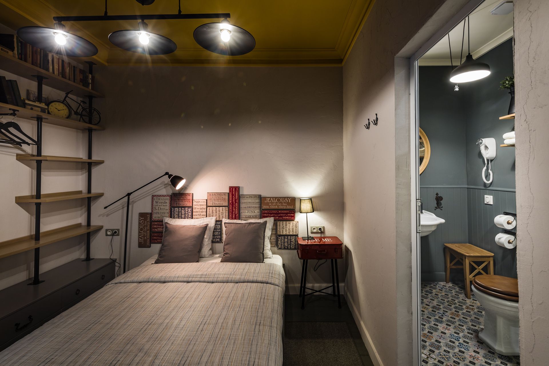 A bedroom with a bed, shelves, a desk, and a bathroom with a toilet and sink.