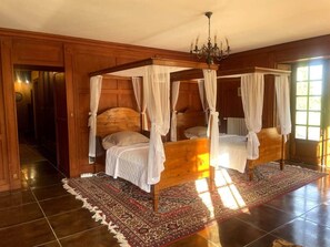 Un lit à baldaquin à quatre poteaux, agrémenté de draps blancs, d’un lustre et d’un tapis à motifs, dans une pièce en bois dotée de grandes fenêtres.