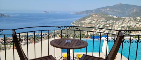 A balcony with two wooden chairs and a small table, overlooking a pool and a coastal town.