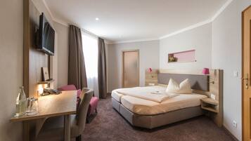 Een hotelkamer met een bed, nachtkastje, televisie, bureau, stoel en een klein plankje met een plant.