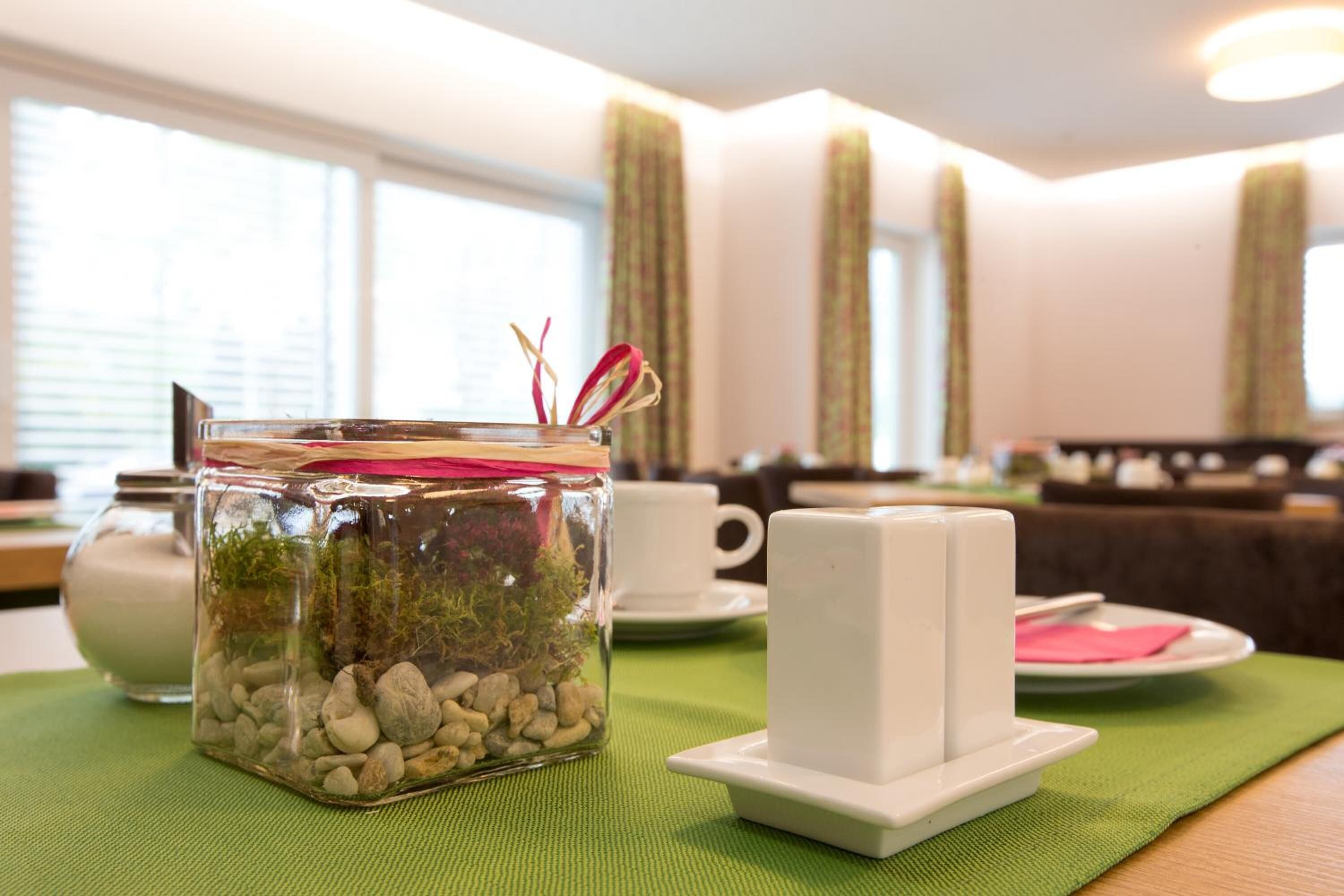 Un bocal en verre contenant des pierres et de la mousse, une tasse blanche et un ensemble salière et poivrière, le tout posé sur une nappe verte dans un coin repas, avec une fenêtre et des rideaux en arrière-plan.