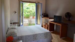 A bedroom with a bed, a desk, a chair, a TV, and a balcony with a view of greenery. - Pousada Santa Maria (Tiradentes)