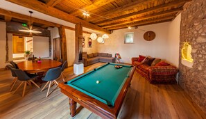 A cozy room with a pool table, wooden ceiling beams, stone walls, and a dining area with chairs and a table.