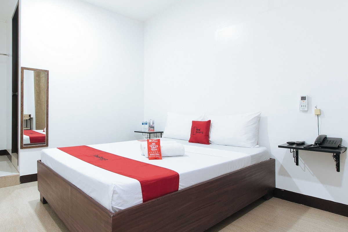 Ein Hotelzimmer mit einem großen Bett, einem Nachttisch mit Telefon und kleiner Lampe, einem Spiegel und einem roten Kopfkissen.