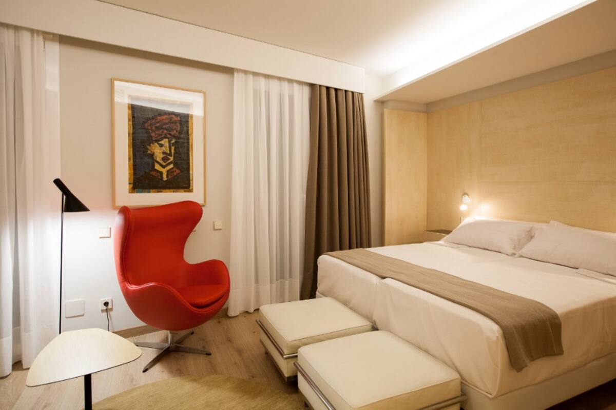 Una habitación de hotel moderna con una cama grande, una silla roja, una mesita de noche y una lámpara de pared.