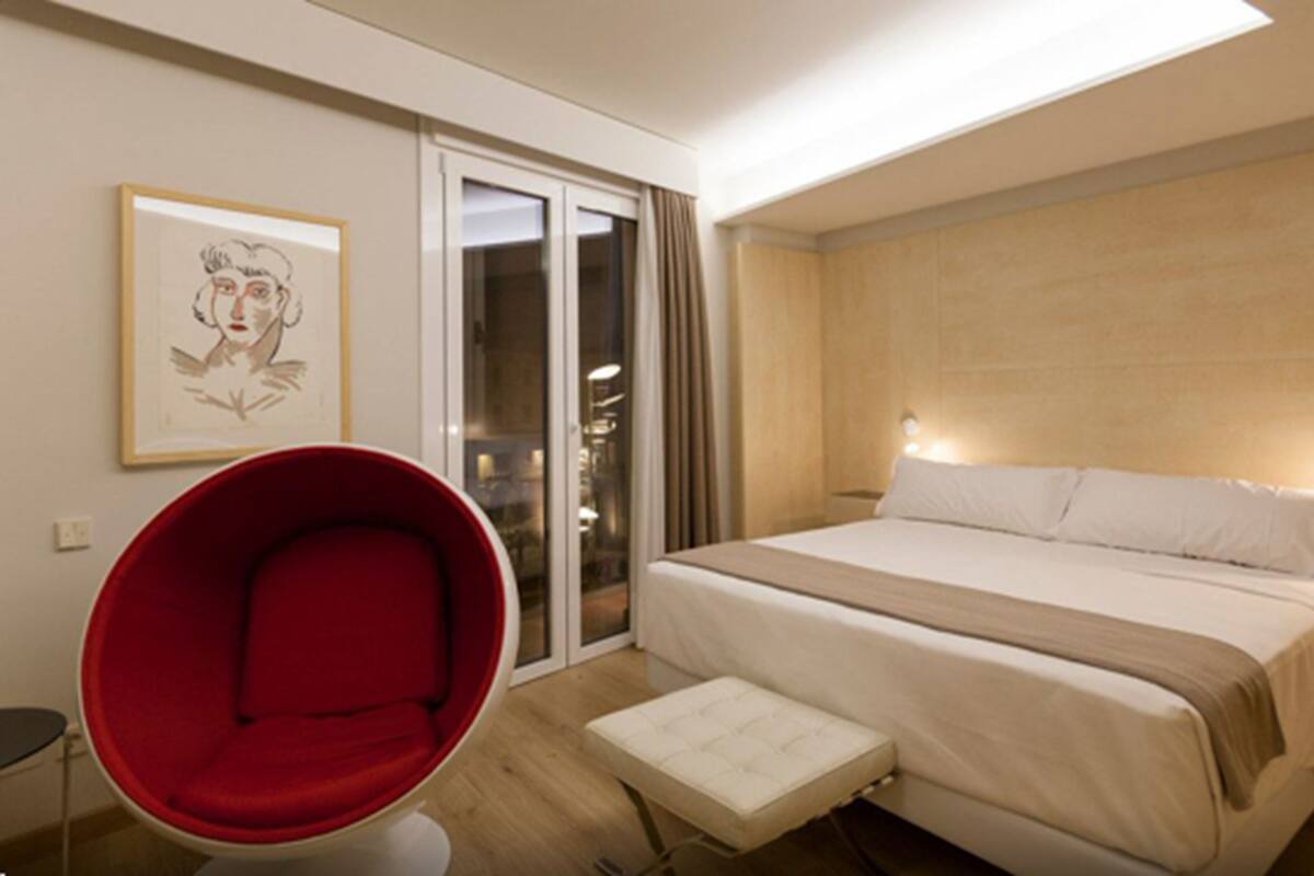 Habitación de hotel moderna con una cama grande, una silla roja, una mesita y un vidrio corredizo que da a un balcón.